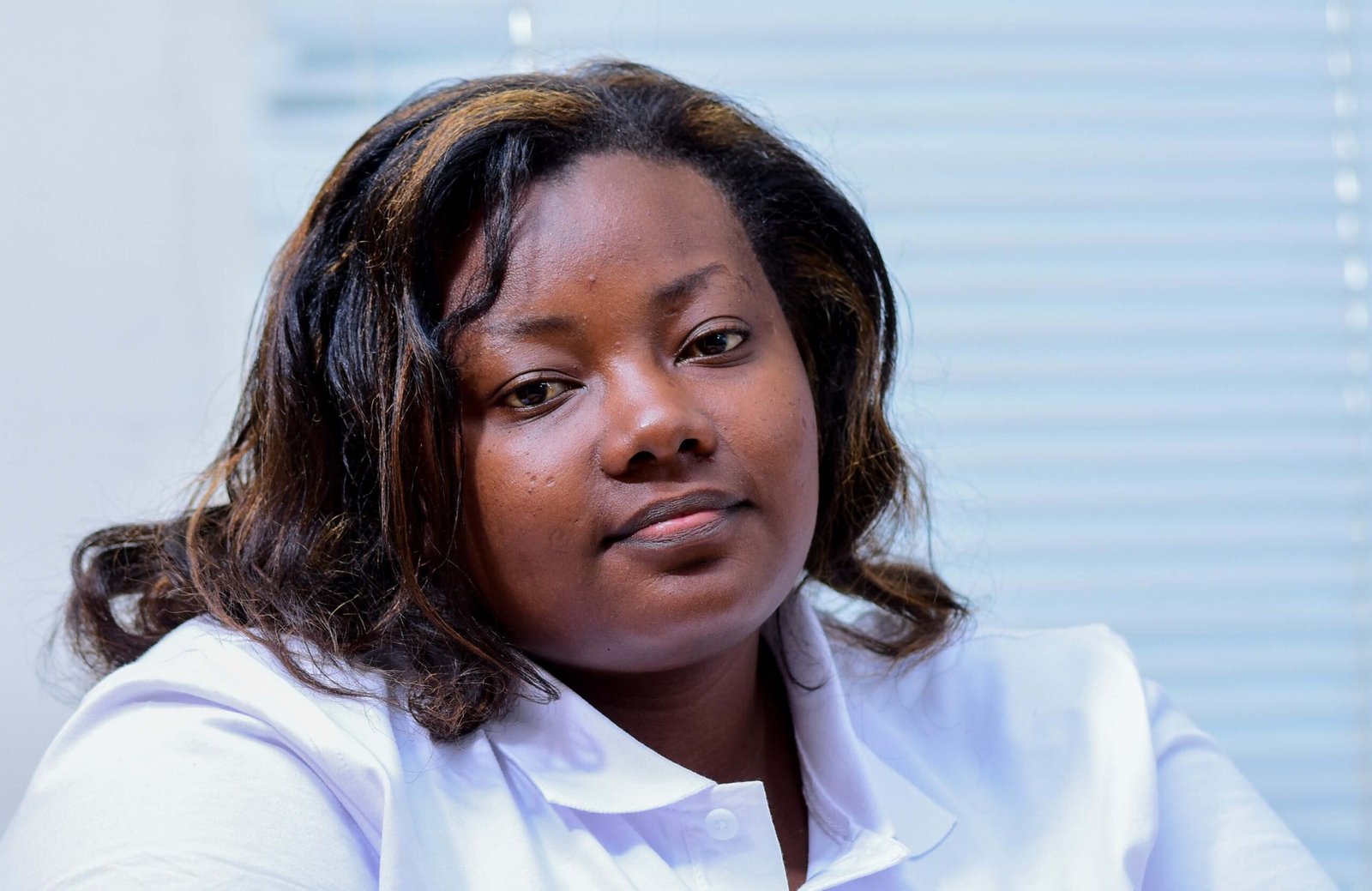Portrait of a Black woman with shoulder-length wavy hair, wearing a light-colored collared shirt, looking calmly at the camera against a softly blurred indoor background.