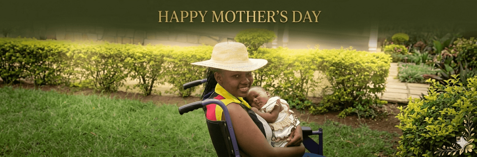 A wide panoramic image with a 3:1 aspect ratio featuring a smiling woman in a straw hat sitting in a purple wheelchair, cradling a sleeping infant in her arms. The scene is set outdoors in a lush green garden with manicured hedges and a stone path in the background. Across the top of the image, the text "HAPPY MOTHER'S DAY" is written in a classic gold font against a dark, blurred green banner. In the bottom right corner, there is a delicate gold floral accent.
