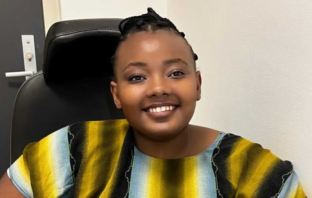 A smiling young woman with braided hair sits indoors in a black chair, wearing a patterned yellow, black, and blue outfit. She looks warmly toward the camera, creating a calm and confident portrait.
